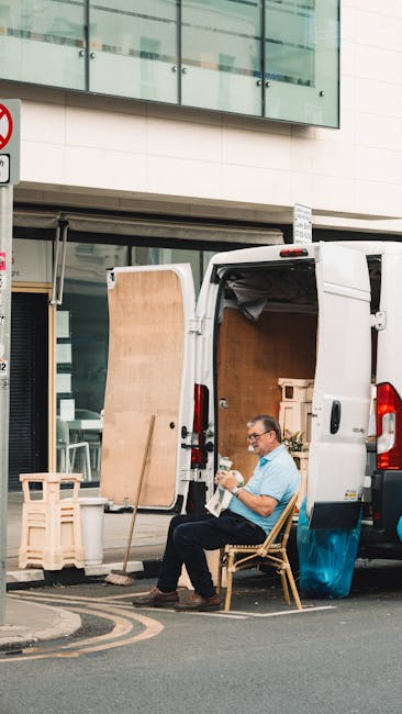 A man wearing a white t-shirt and jeans is standing on the curbside outside a residential property, loading cardboard boxes into the open rear doors of a white moving van. The boxes vary in size and a