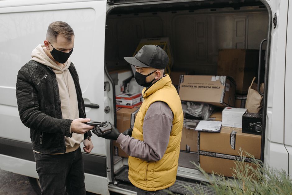 A man wearing a beige knitted beanie, green puffer vest, and dark blue sports jacket with white stripe detailing is sitting on the pavement in front of an open van during a home relocation process. Th