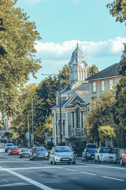 A busy street scene in Selsdon showing several parked and moving cars along a paved road. In the background, there is a historic building with a clock tower, featuring a pointed roof, arched windows, and decorative stonework. Tall trees with green foliage line the street, casting shade over the parked vehicles and buildings. The sky is clear with some white clouds, and the area appears calm and well-maintained. This setting reflects the environment where house removals and furniture transport services by Man and Van Selsdon are often carried out, involving logistics of loading and unloading within residential and commercial locations around Selsdon. The scene emphasizes the typical urban street environment suitable for home relocation activities, vehicle loading, and moving service operations.