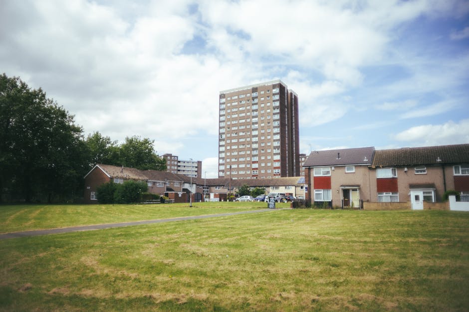 A residential area in Selsdon featuring a large, multi-storey apartment building with a brown brick exterior and numerous windows, situated behind a row of smaller, single-story houses with tiled roofs and light-colored walls. The houses are set along a paved pathway, with a well-maintained grassy lawn in the foreground. Several parked cars are visible near the houses, and the sky above is partly cloudy with patches of blue. The scene depicts an exterior view during daylight hours, with natural lighting highlighting the urban environment suitable for house removals, furniture transport, and packing and moving processes, as managed by Man and Van Selsdon.