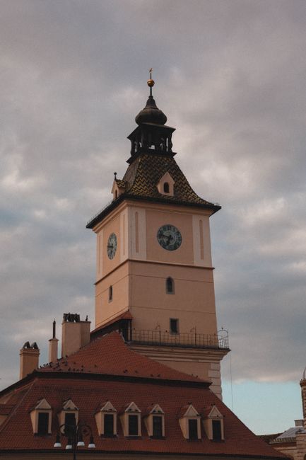 A busy street scene in Selsdon showing several parked and moving cars along a paved road. In the background, there is a historic building with a clock tower, featuring a pointed roof, arched windows, and decorative stonework. Tall trees with green foliage line the street, casting shade over the parked vehicles and buildings. The sky is clear with some white clouds, and the area appears calm and well-maintained. This setting reflects the environment where house removals and furniture transport services by Man and Van Selsdon are often carried out, involving logistics of loading and unloading within residential and commercial locations around Selsdon. The scene emphasizes the typical urban street environment suitable for home relocation activities, vehicle loading, and moving service operations.