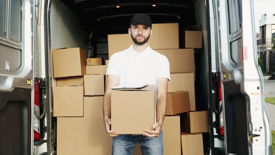 A young man with short dark hair, wearing a white t-shirt and jeans, is standing on the pavement next to a white cargo van during a home relocation process. He is holding a cardboard box and appears t