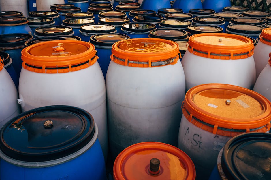 A close-up view of numerous large plastic drums and barrels arranged outdoors, with some white barrels featuring orange lids and others blue barrels with black or orange lids, in a setting that appears to be related to home relocation or storage. The barrels, used for transporting or storing materials, are positioned on a paved surface and are arranged in rows, with some barrels overlapping or partially obscured by others. The lighting suggests daylight, and the barrels are clean and sealed, indicating proper handling and packing during moving or logistical operations. Man and Van Selsdon, a professional removal service, would typically handle the transport and safe storage of such items during house moves and relocation projects, ensuring compliance with safety and waste management regulations, especially when dealing with hazardous waste or materials requiring careful handling during the loading process.