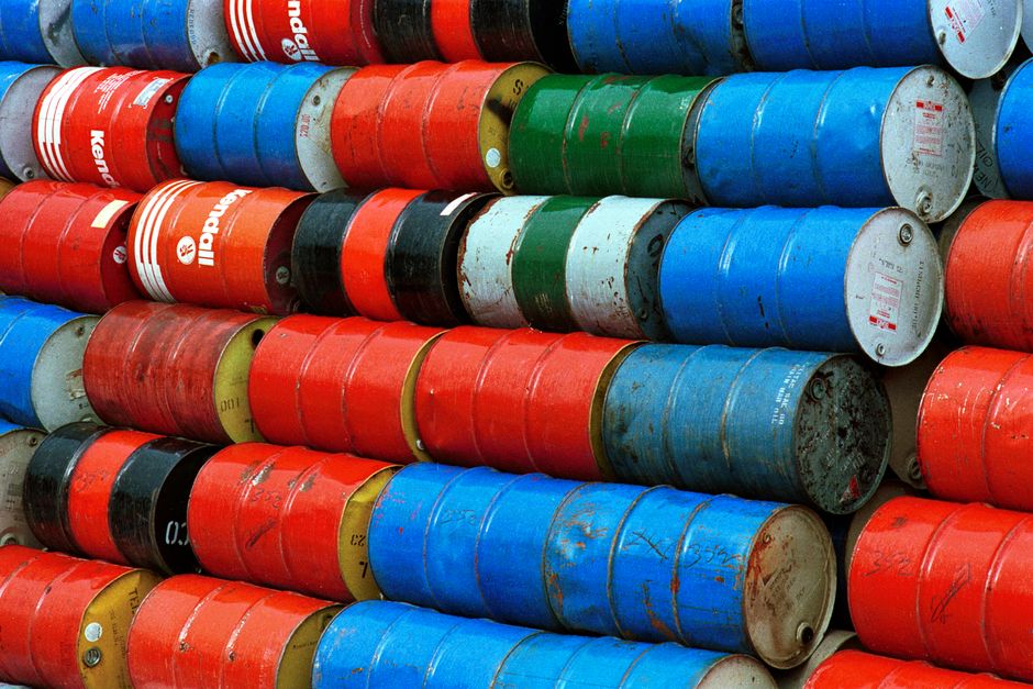 A large collection of cylindrical metal drums stacked horizontally and vertically in an outdoor area, with colors including red, blue, green, yellow, and black. Many drums show signs of rust, dirt, and paint chipping, indicating wear and age. They are arranged in a closely packed pattern, covering most of the image, with some resting on wooden pallets and others directly on the ground. The background features more drums similarly stacked, suggesting a storage or disposal site for hazardous waste, relevant to house removals and relocation services. The image depicts an industrial environment, possibly near a warehouse or waste management facility, emphasizing the importance of proper handling and disposal during home relocation or furniture transport processes, as managed by companies like Man and Van Selsdon.