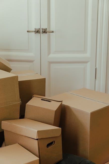 Inside a residential room with a closed white door featuring silver hinges, several cardboard moving boxes of various sizes are stacked near the door. The boxes include a small rectangular box with a handle, a medium-sized box with a lid, and larger boxes possibly containing household items. The boxes are made of brown corrugated cardboard and appear to be packed for a home relocation. The room has a light-colored wall and door trim, and the floor is not visible. This scene depicts the packing phase of a house move, typical of furniture transport and moving services provided by companies like Man and Van Selsdon. The focus is on the organized stacking of packing materials prepared for loading onto a van, supporting efficient and careful furniture and item transport during the home removal process.