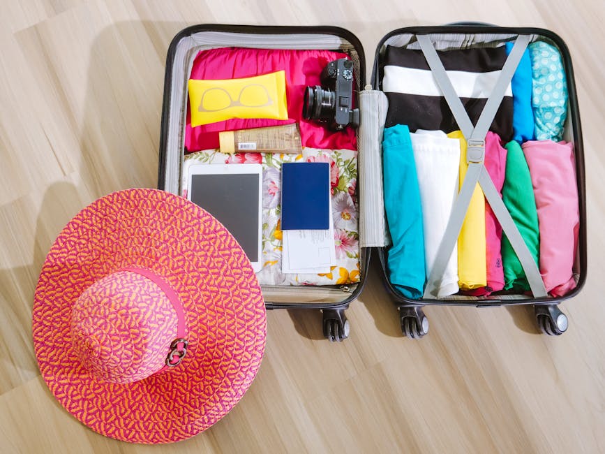 An open black hard-shell suitcase on a light wooden floor, containing neatly packed clothing in various colors, including teal, pink, yellow, and black-and-white striped items, secured with grey straps inside, alongside a pink patterned sunhat placed nearby. The suitcase lid displays packing accessories such as a black camera, a smartphone, a tablet, a small bottle, and a floral-patterned cloth. The setting appears to be in a residential interior, illustrating the packing process for a home relocation or furniture transport, as part of the services offered by Man and Van Selsdon during a house move or flat packing and moving process.