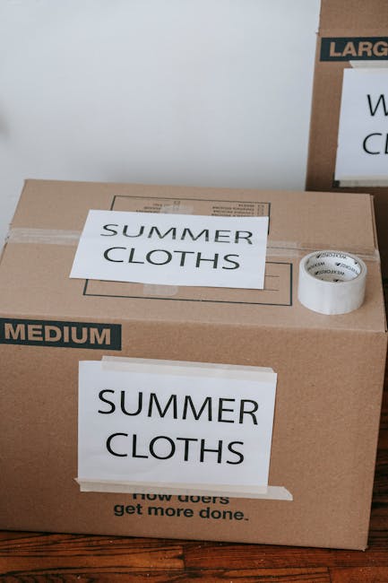 Inside a residential room with a closed white door featuring silver hinges, several cardboard moving boxes of various sizes are stacked near the door. The boxes include a small rectangular box with a handle, a medium-sized box with a lid, and larger boxes possibly containing household items. The boxes are made of brown corrugated cardboard and appear to be packed for a home relocation. The room has a light-colored wall and door trim, and the floor is not visible. This scene depicts the packing phase of a house move, typical of furniture transport and moving services provided by companies like Man and Van Selsdon. The focus is on the organized stacking of packing materials prepared for loading onto a van, supporting efficient and careful furniture and item transport during the home removal process.