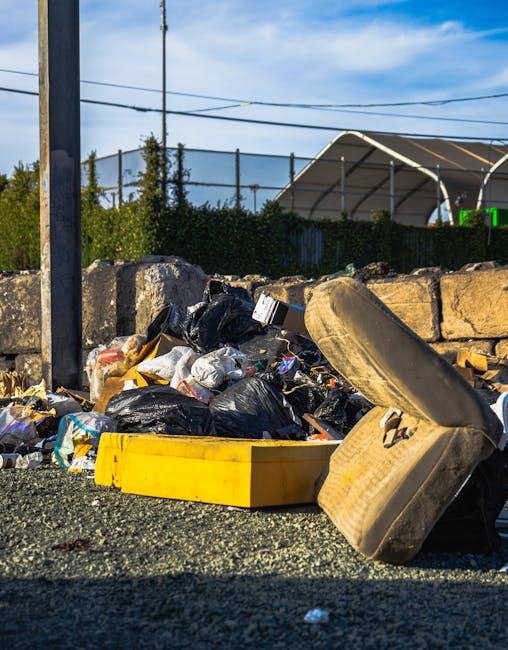 A pile of discarded waste including black plastic rubbish bags, a yellow plastic container, and an old, worn-out car seat lying on the pavement outside a property, with a stone wall and a fence in the background. The waste is situated near a metal pole and under power lines, indicating disposal of bulky or unwanted items following a home relocation or moving process, as handled by a professional removals service such as Man and Van Selsdon. The scene is outdoors, during daylight hours, with a partly cloudy sky overhead, and the area appears to be an informal waste disposal spot close to a residential or commercial area.