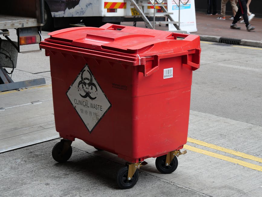 A red plastic clinical waste bin with four wheels is positioned outdoors on a concrete surface near the rear of a vehicle, likely used for home relocation or waste disposal following a move. The bin features a lid with a slightly raised curvature and a biohazard symbol with the label 'Clinical Waste' affixed to the front. Surrounding the bin are elements of an urban environment, including a curb, double yellow lines, and a nearby sidewalk with pedestrians visible in the background. The area appears well-lit, indicating daytime, and the presence of packaging materials or furniture is not visible, suggesting the bin is designated for waste collection during moving processes. Man and Van Selsdon, a specialist in removals and furniture transport, may handle or advise on safe disposal of waste after a home relocation, aligning with the context of this image.