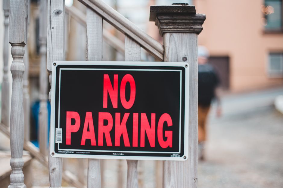 A close-up of a rectangular 'No Parking' sign attached to a wooden railing outside a residential property. The sign features a black background with bold, uppercase red letters spelling 'NO PARKING,' framed by a thin white border. Behind the sign, part of a wooden staircase with vertical spindles and a newel post is visible, leading to the entrance of a house with a pinkish exterior wall and a window. In the background, a blurred view of parked vehicles and a street suggests an urban or suburban setting. The placement of the sign indicates parking restrictions relevant to house removals or moving logistics managed by Man and Van Selsdon, supporting the context of local relocation services and parking regulations in Selsdon, especially during the loading process of furniture and moving boxes.
