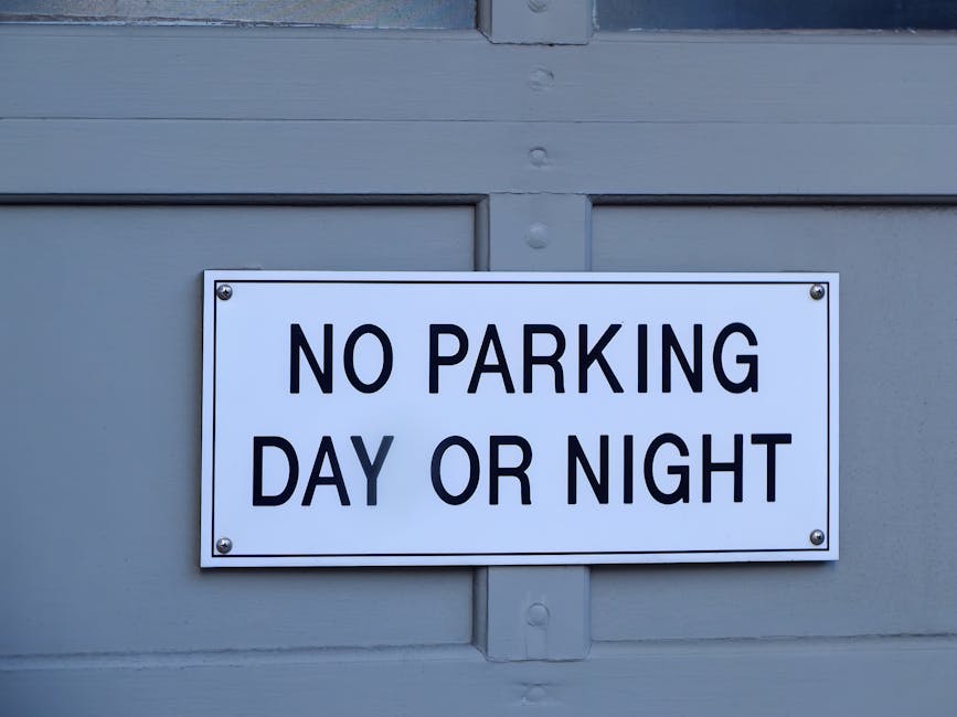 A rectangular white sign with bold black lettering reading 'NO PARKING DAY OR NIGHT' affixed to a light grey wooden door or wall. The sign is secured at each corner with metal screws, and the surface of the sign is smooth and clean. The background consists of horizontal wooden panels painted in a neutral grey tone, with visible seams and wood grain texture. The image appears to have been taken during daylight hours, as natural light illuminates the scene evenly. The sign likely indicates parking restrictions in the area, relevant to house removals or moving services in Selsdon, as seen on the webpage for Man and Van Selsdon, which specialises in removals and furniture transport.