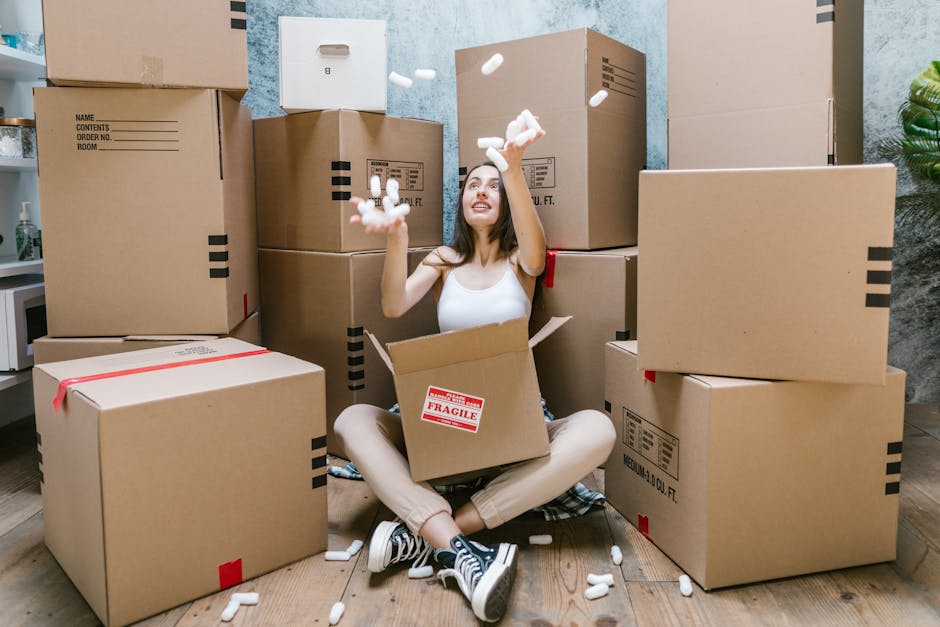 A woman sitting on a wooden floor inside a room, surrounded by numerous large cardboard moving boxes, some labeled with red 'Fragile' stickers and others with printed handling instructions. She is smi