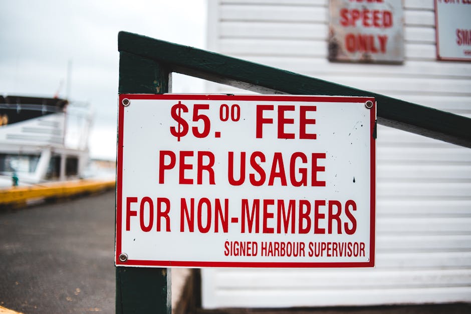 A rectangular white sign with bold red lettering displaying a fee of $5.00 per usage for non-members, attached to a dark green metal railing with four silver screws. The sign is situated outdoors near