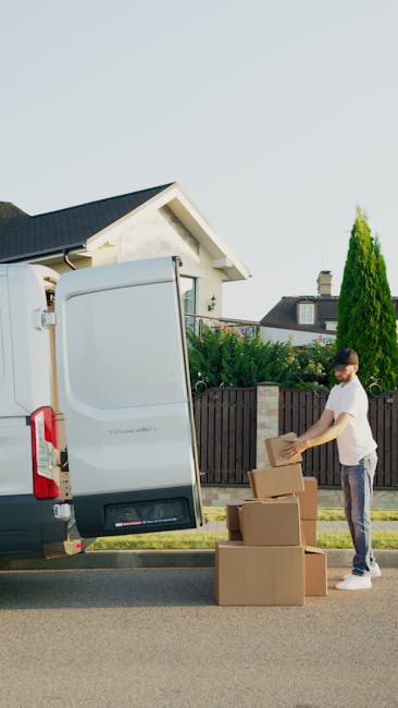 A man with short dark hair, wearing a white t-shirt and jeans, is standing on the pavement next to a large white moving van with its rear doors open. He is loading cardboard boxes of various sizes onto the vehicle, carefully stacking them on the ground. Inside the van, visible are wrapped furniture items and packed cardboard boxes, indicating a home relocation. The loading process occurs outdoors in a suburban residential area, with a detached house featuring a gabled roof and a chimney in the background, along with neatly maintained bushes and trees. The scene is well-lit with natural daylight, and the surroundings include a wooden fence and neighboring houses, emphasizing the typical environment for house removals and furniture transport handled by [COMPANY_NAME], who offer professional removal and moving services in Selsdon.